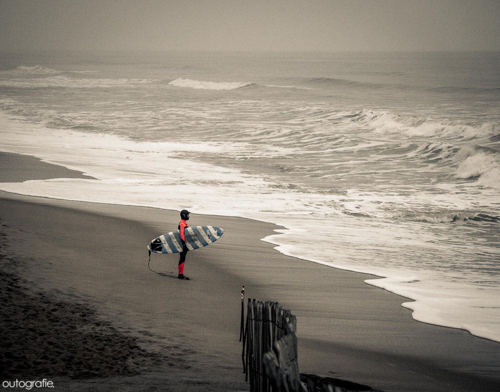 Kleiner Surfer mit warmem Wetsuit, Wetsuit Haube, Wetsuit Schuhen und Wetsuit Handschuhen schaut auf die kalten Winter Wellen. 