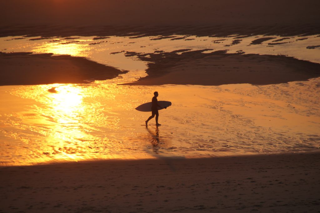 Surfer läuft durch den Sonnenuntergang. Nach einer langen Surfsession lockt das wohlverdiente Feierabendbier. Doch Alkoholkonsum könnte Sodbrennen beim Surfen verursachen.