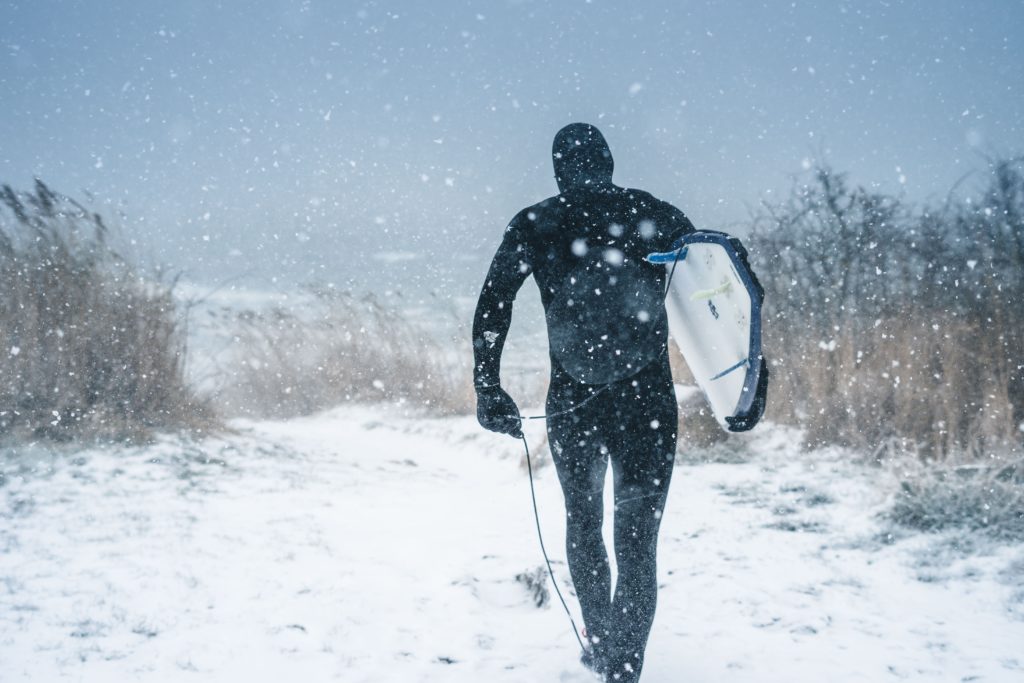 Ein Surfer läuft mit seinem Board durch einen Schneesturm. Vorbeugung und Früherkennung des Surfers Ear sind vor allem für Kaltwasser Surfer relevant.
